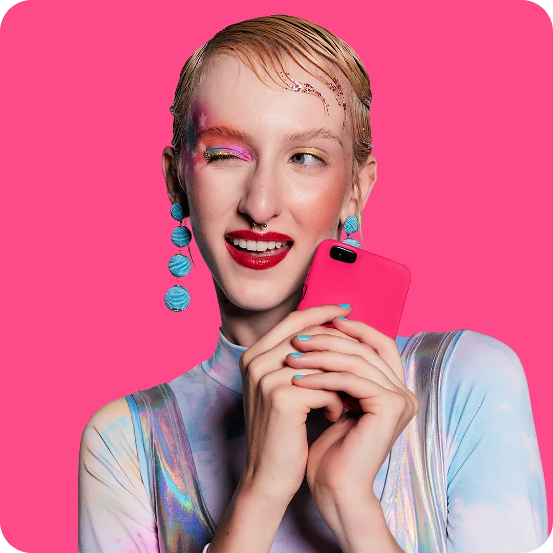 A person with short blonde hair, bold makeup, and blue earrings smiles and winks while holding a pink phone, wearing a pastel outfit against a bright pink background.
