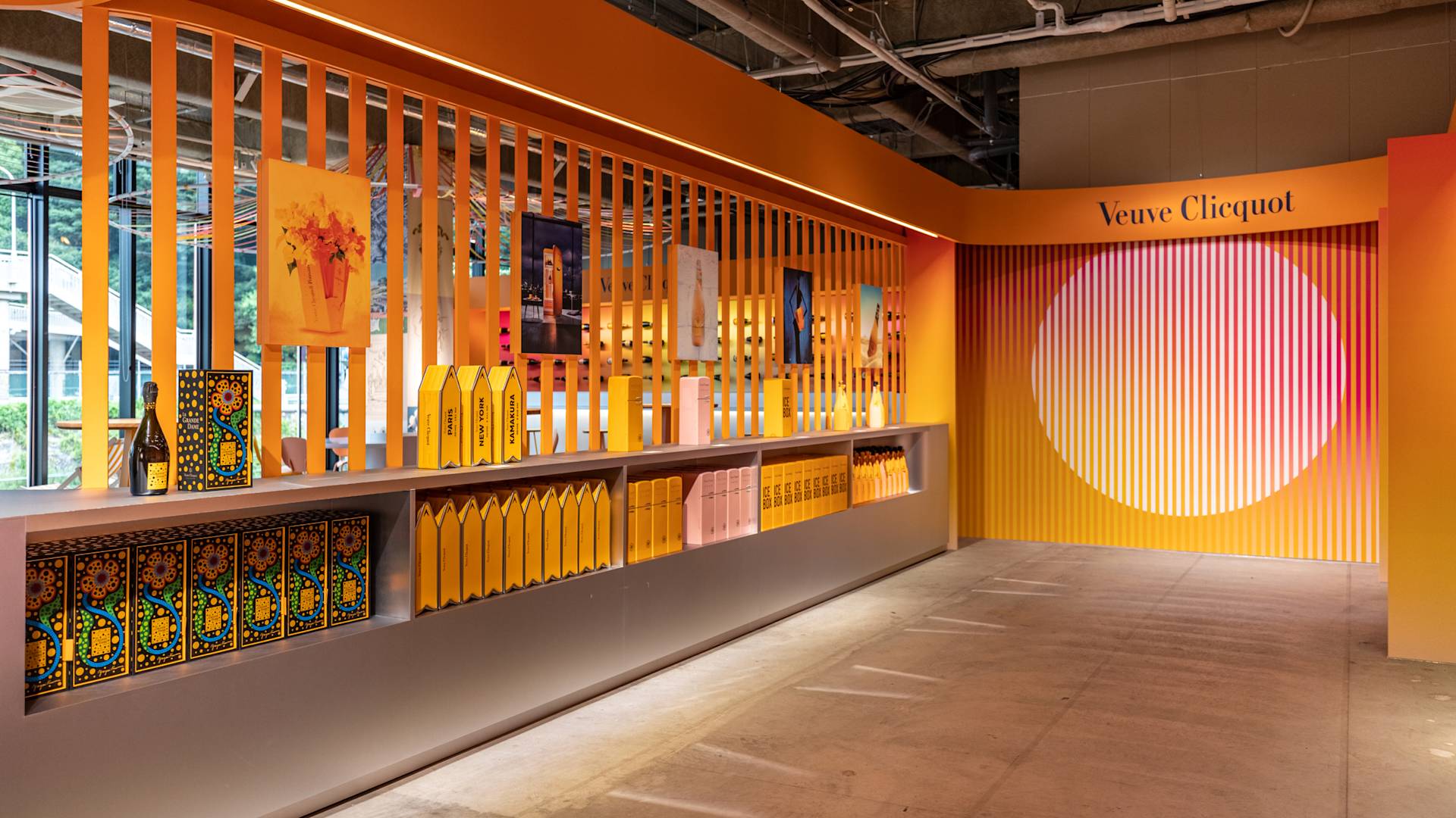 A modern, brightly lit Veuve Clicquot display features yellow and orange packaging, artwork, and bottles arranged neatly on shelves, with bold striped panels and a large circular design on the wall.