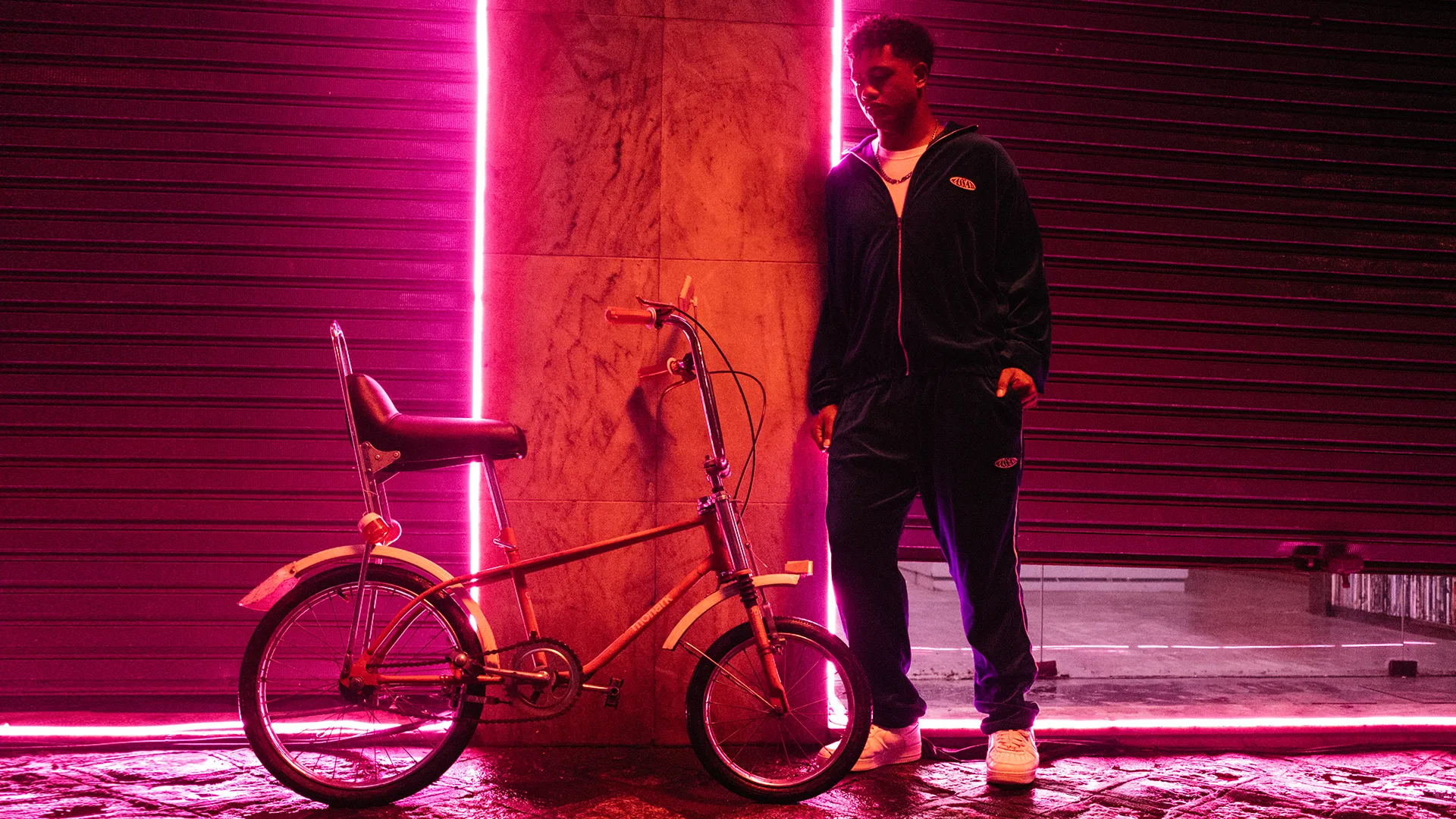 A person in a dark tracksuit stands next to a vintage bicycle in front of a wall with bright pink neon lighting, casting a colorful glow over the scene at night.