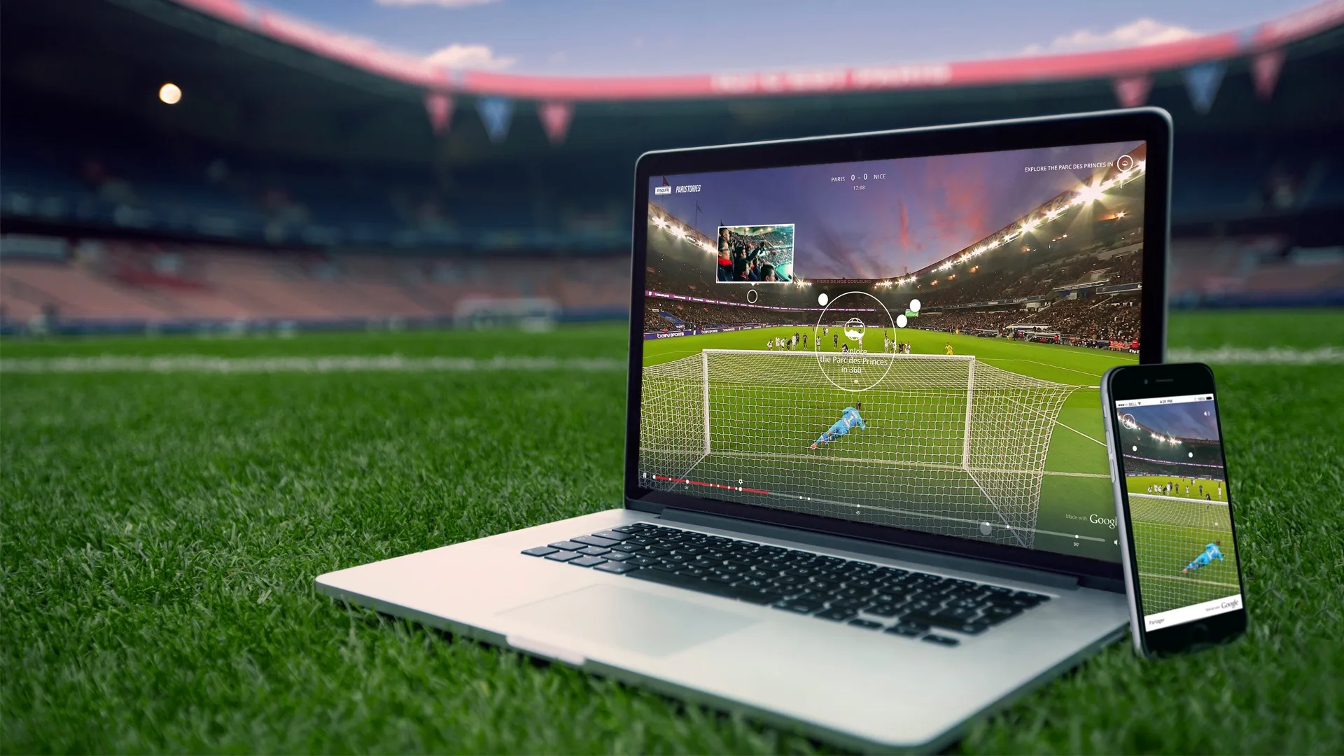 A laptop and smartphone on a soccer field display a live soccer match, showing a goal attempt from behind the goal line, with the stadium visible in the background.
