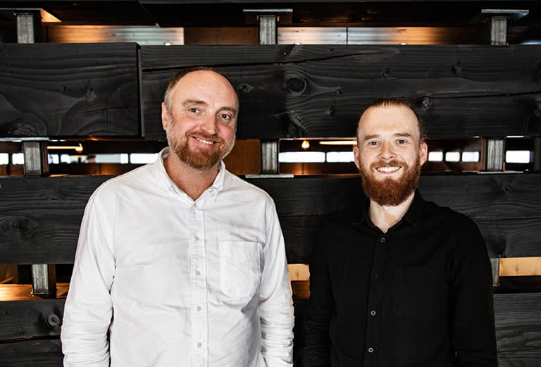 Two men with beards stand side by side, smiling at the camera. One wears a white button-up shirt and the other wears a black shirt. They are indoors in front of a dark wooden backdrop with horizontal panels.