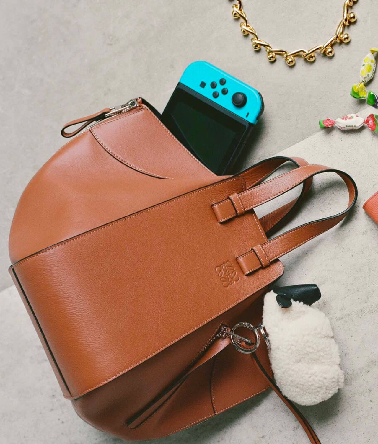 A brown leather handbag with a blue Nintendo Switch partially sticking out, a white fluffy keychain attached, a gold necklace, and colorful wrapped candies nearby on a light surface.