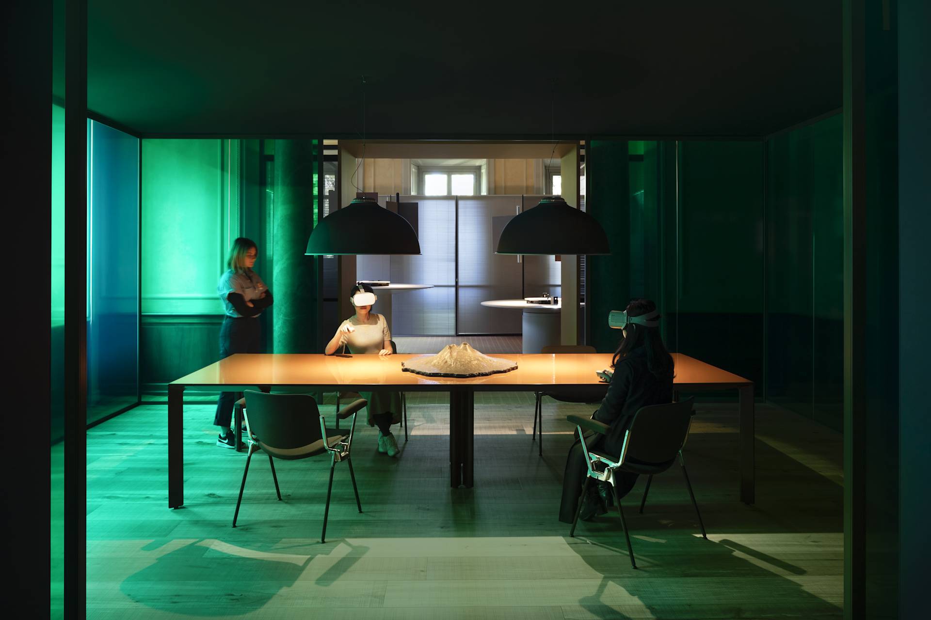 Three people in a dim, modern room sit at a large table; two wear VR headsets, while one stands nearby. A sculptural object is on the table under two hanging lamps, with green-tinted glass walls surrounding them.