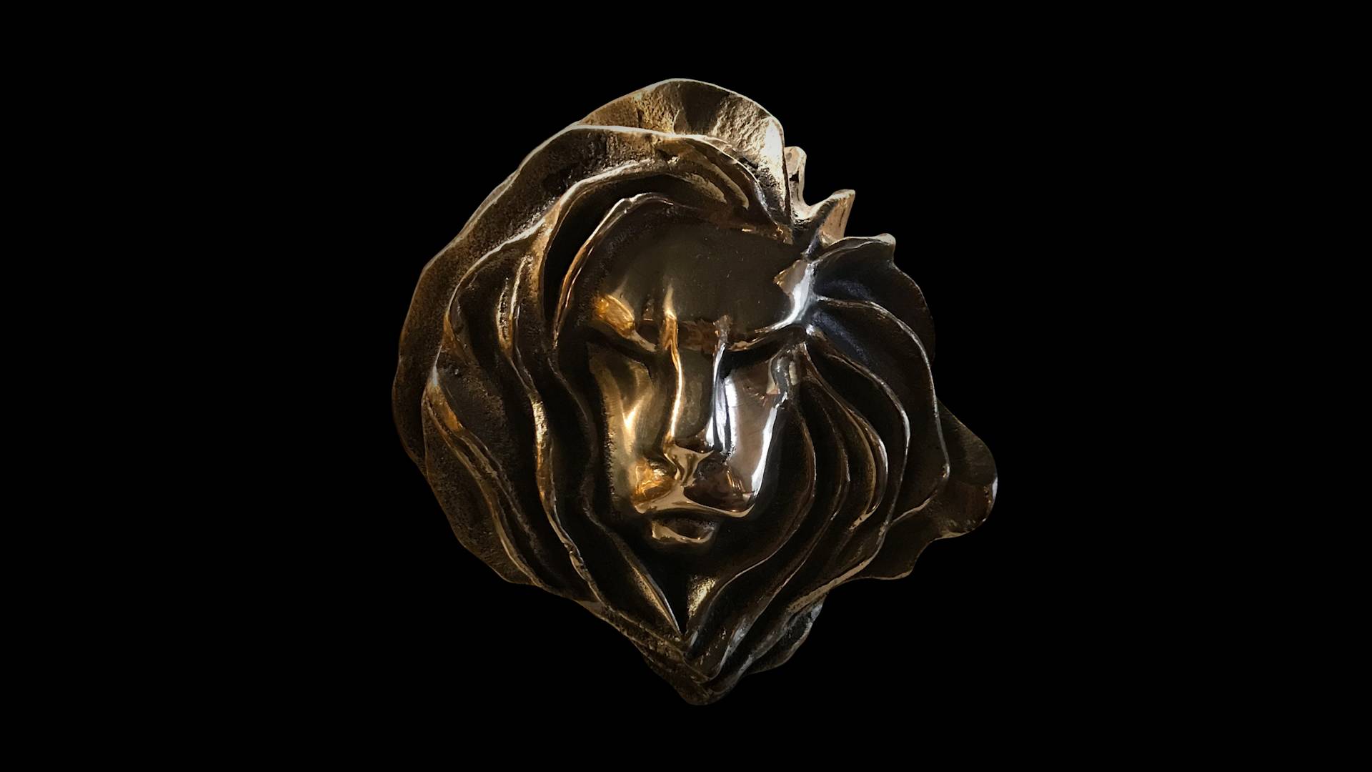 A bronze sculpture of a lion’s head with stylized, flowing mane details, shown against a black background.