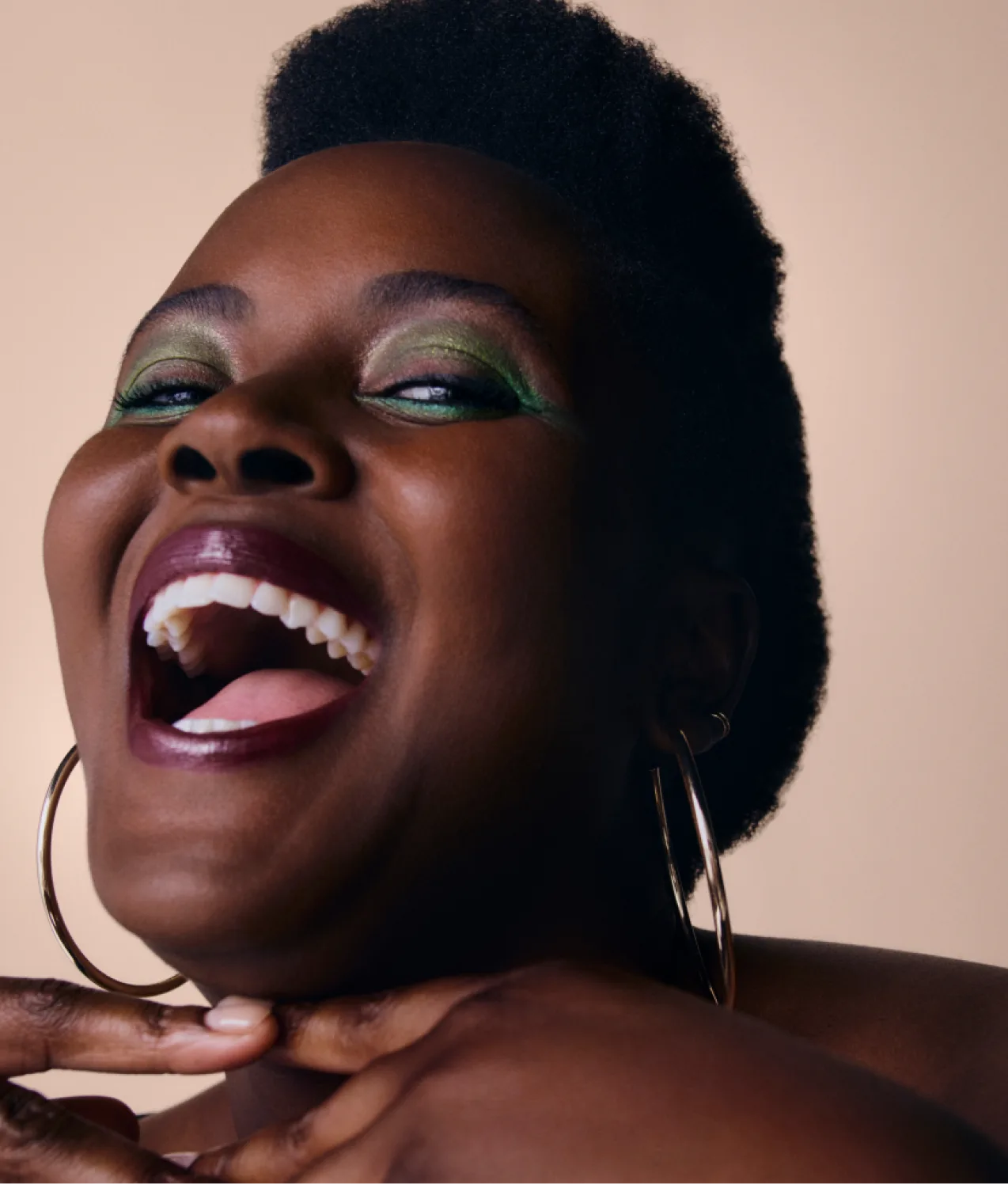 A joyful woman with dark skin smiles widely, showing her teeth. She has green eye shadow, dark lipstick, large hoop earrings, and short natural hair. Her fingers are gently touching her chin against a neutral background.