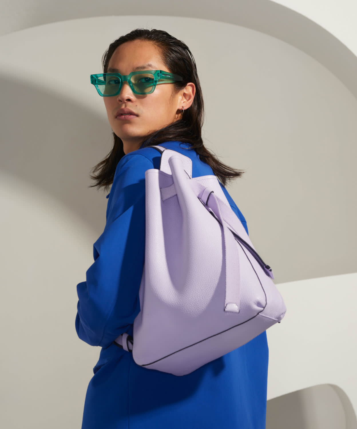 A person wearing a bright blue top and green translucent glasses looks over their shoulder, modeling a light purple backpack against a minimalist, curved white background.