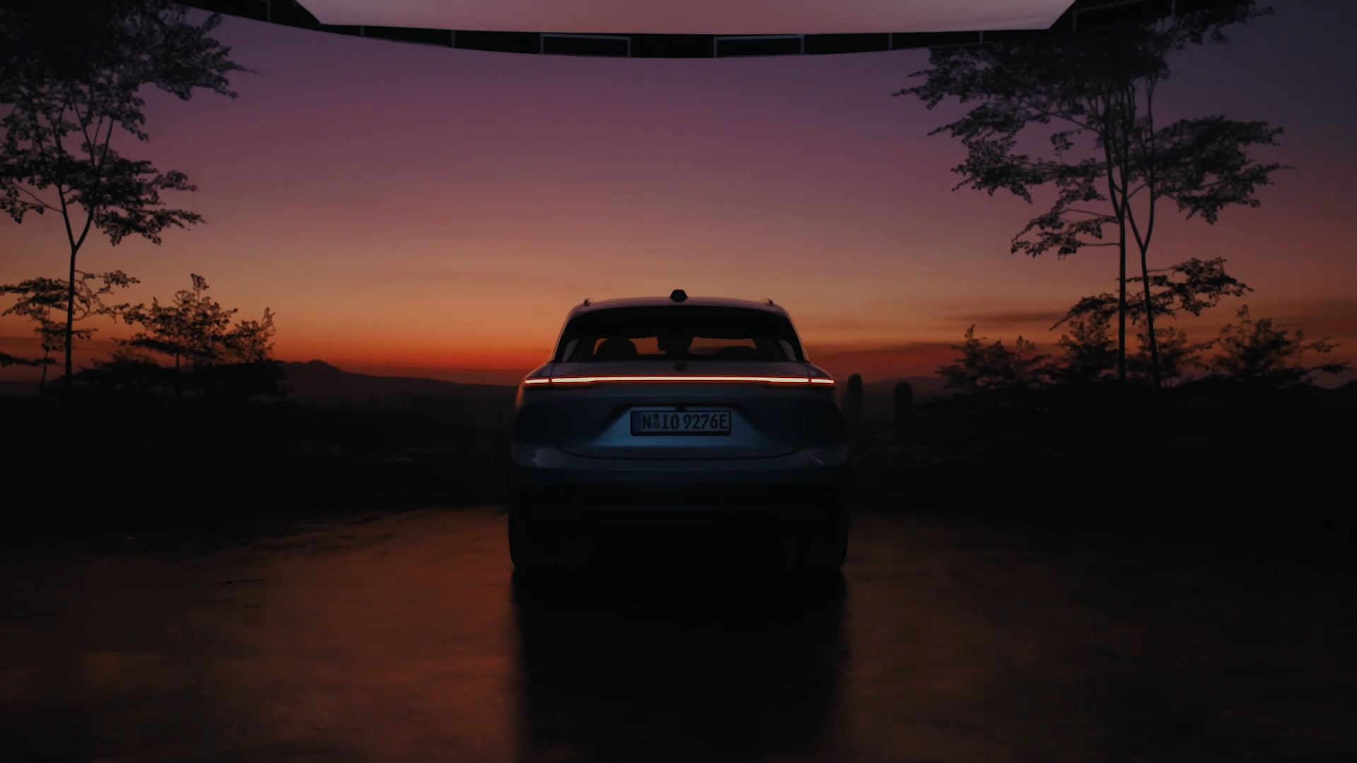 A car is parked outdoors at dusk, viewed from behind, with a vivid orange and purple sunset sky in the background and silhouettes of trees on each side.