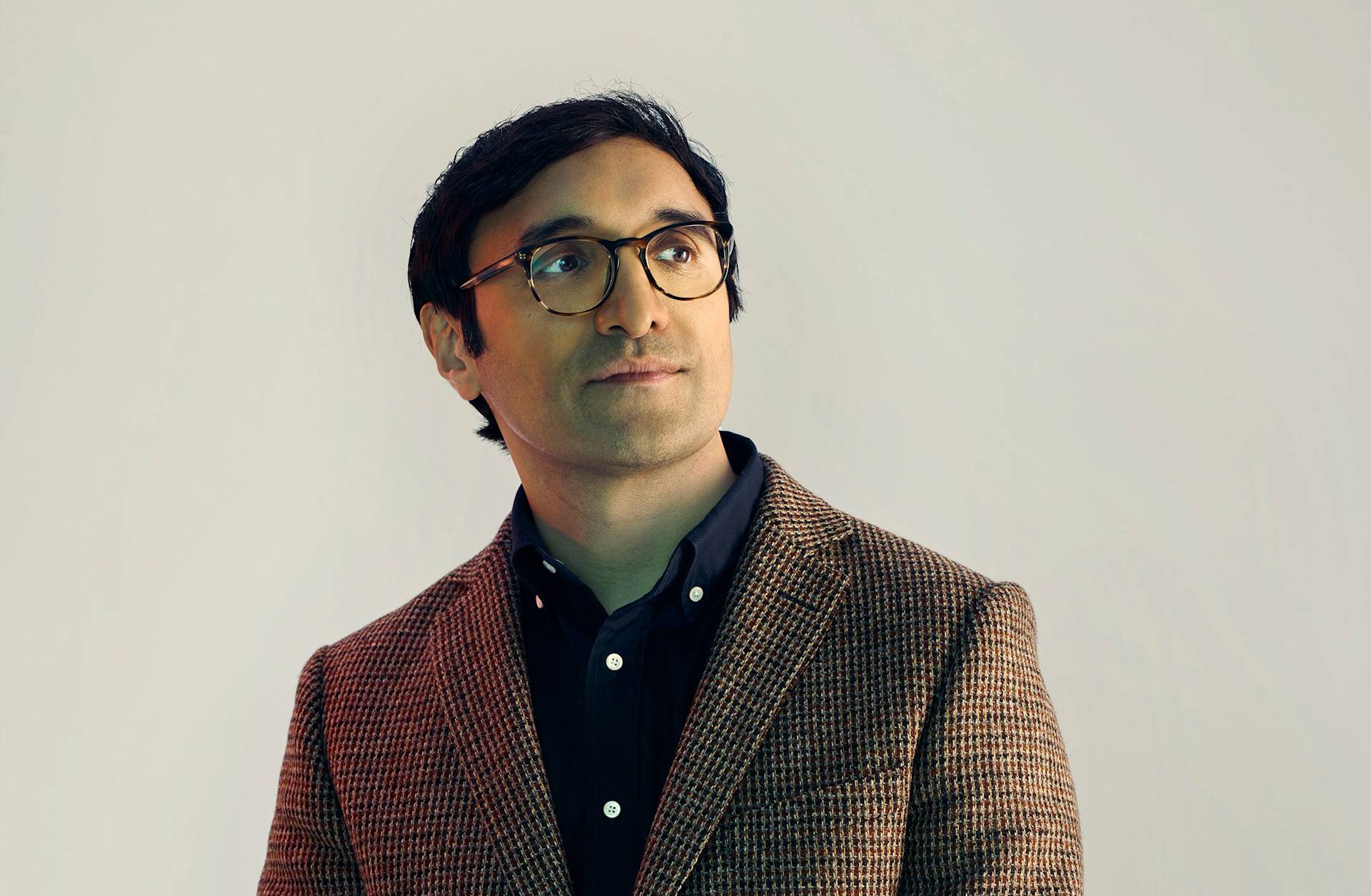 A man with short dark hair and glasses wears a brown checkered blazer over a dark shirt, looking slightly upward and to the side against a plain light background.