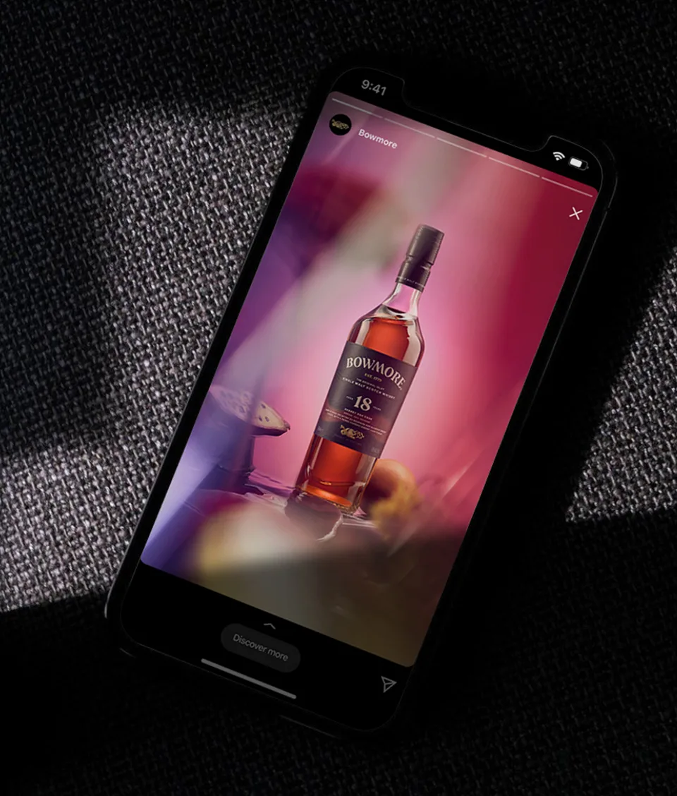 A smartphone displays an Instagram story featuring a Bowmore 18-year-old whisky bottle set against a blurred, pink and purple background, resting on a textured dark fabric surface.