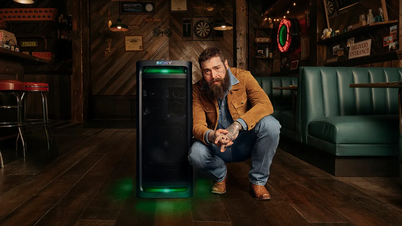 A man with a beard and tattoos crouches next to a large Sony speaker with green lights in a retro-style bar with wooden floors, green booths, and eclectic wall decor.