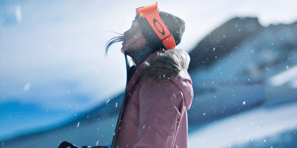 A person in a pink winter coat and orange ski goggles looks up, standing in falling snow with a snowy mountain in the background.