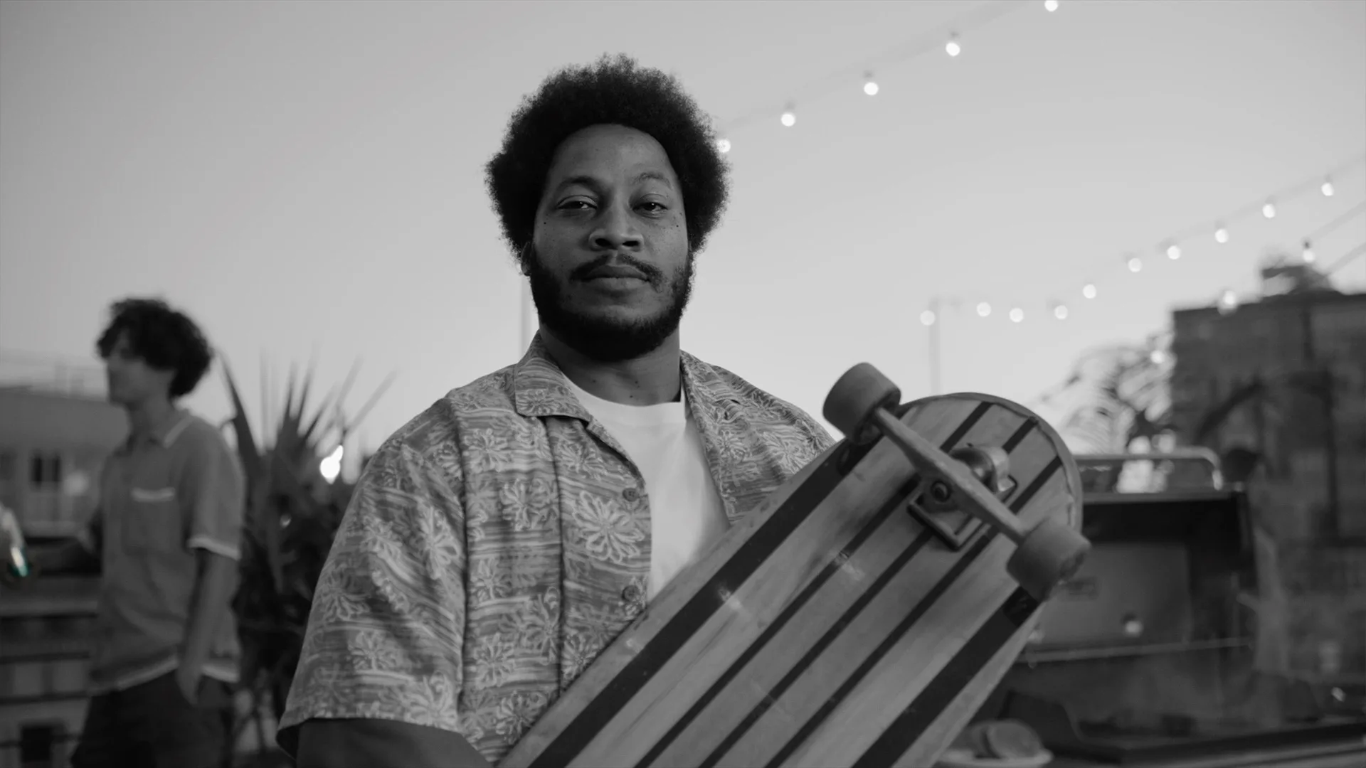A person with an afro hairstyle holds a striped skateboard and looks at the camera. String lights glow above, and another person stands blurred in the background. The setting appears to be an outdoor gathering at dusk.