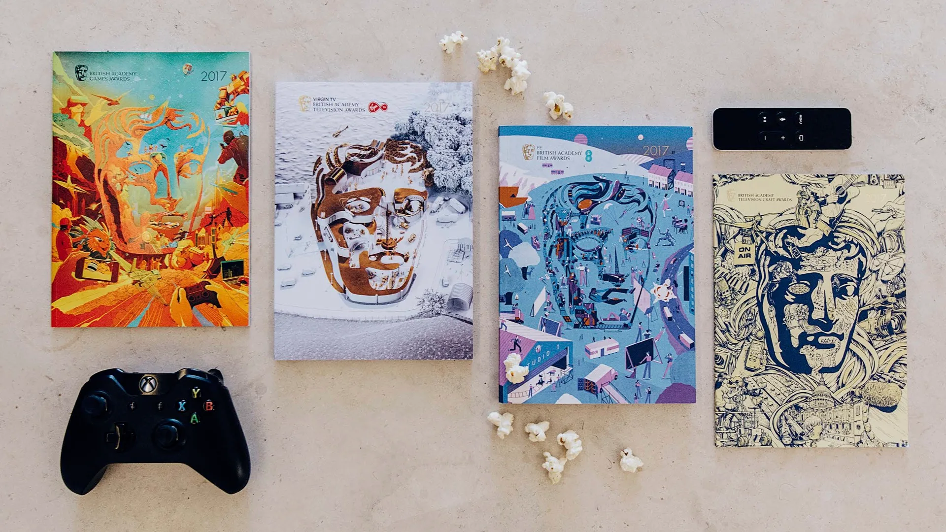 Four illustrated BAFTA booklets for 2017 are arranged in a row on a beige surface, with popcorn pieces scattered around. An Xbox controller and a remote are placed on either side of the booklets.