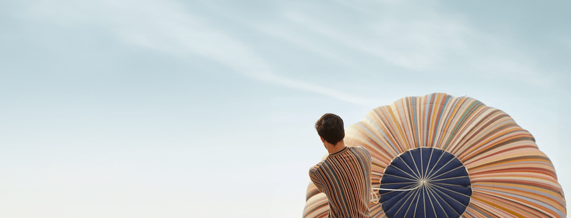 A person in a striped shirt holds the edge of a large, colorful parachute under a clear blue sky with wispy clouds.