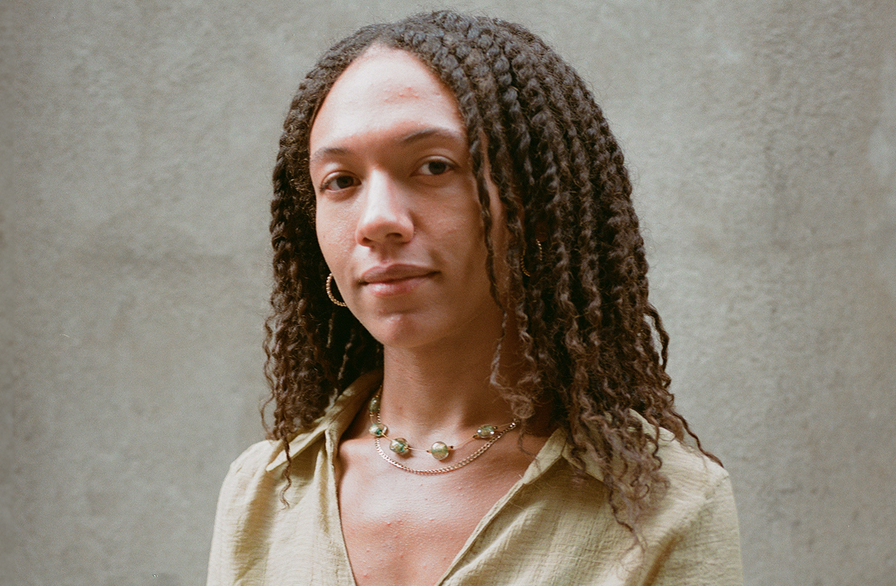 A person with medium-length curly hair, wearing a light-colored shirt, gold hoop earrings, and a beaded necklace, stands in front of a plain, textured background, looking calmly at the camera.