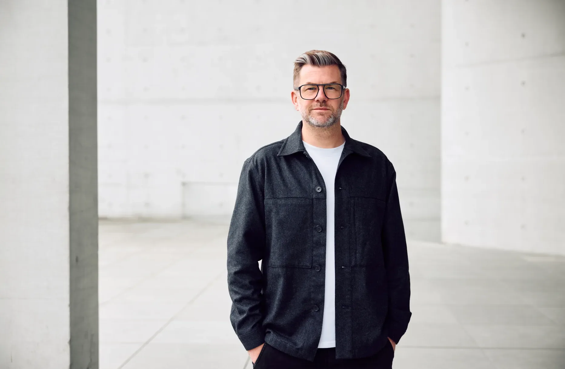 A man with glasses and gray hair, wearing a black jacket over a white shirt, stands with hands in pockets in a modern, minimalistic concrete space.