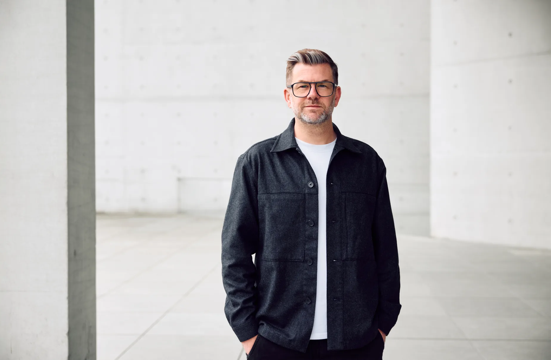 A man with glasses and gray hair, wearing a black jacket over a white shirt, stands with hands in pockets in a modern, minimalistic concrete space.