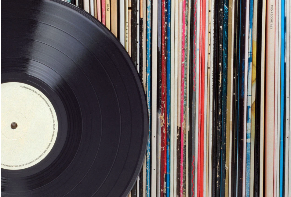 A close-up of a vinyl record partially in view, with its blank label visible, set against a background of vertically stacked vinyl record sleeves showing various colorful spines.