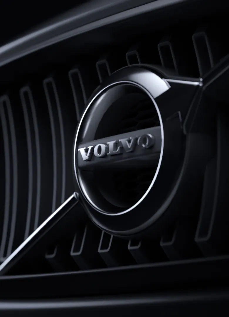 Close-up of a Volvo car grille featuring the chrome Volvo logo in the center, with vertical black grille slats in the background, all in dramatic lighting.