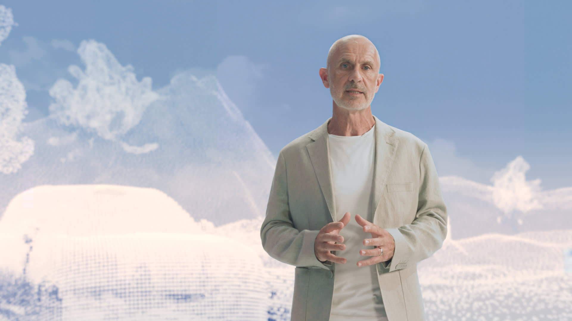 Jim Rowan, wearing a light beige jacket over a white shirt, stands in front of a digital, abstract blue and white background, speaking with his hands clasped.