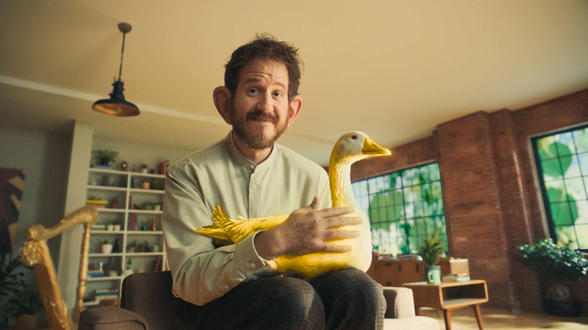A man with a beard and light-colored shirt sits indoors, smiling and holding a large, yellow, duck-shaped object on his lap. The room has shelves, plants, and large windows in the background.