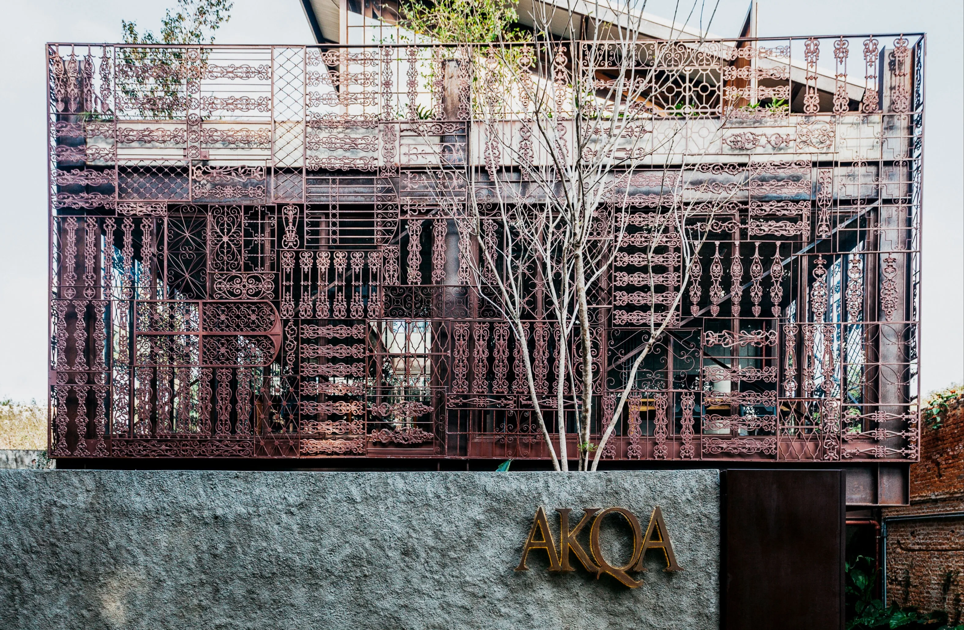 Decorative metal screen with intricate geometric patterns covers the facade of a building. In front, a gray textured wall features the gold signage AKQA. Leafless tree branches rise in front of the structure.