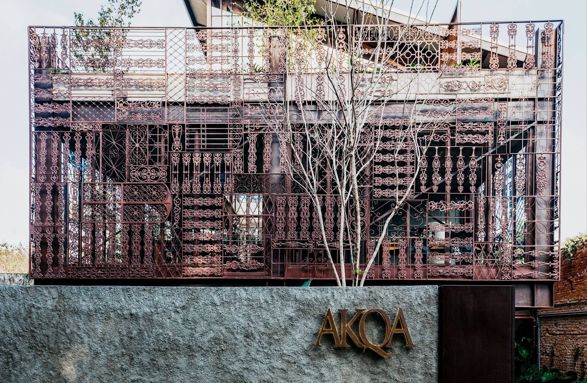 Decorative metal screen with intricate geometric patterns covers the facade of a building. In front, a gray textured wall features the gold signage AKQA. Leafless tree branches rise in front of the structure.