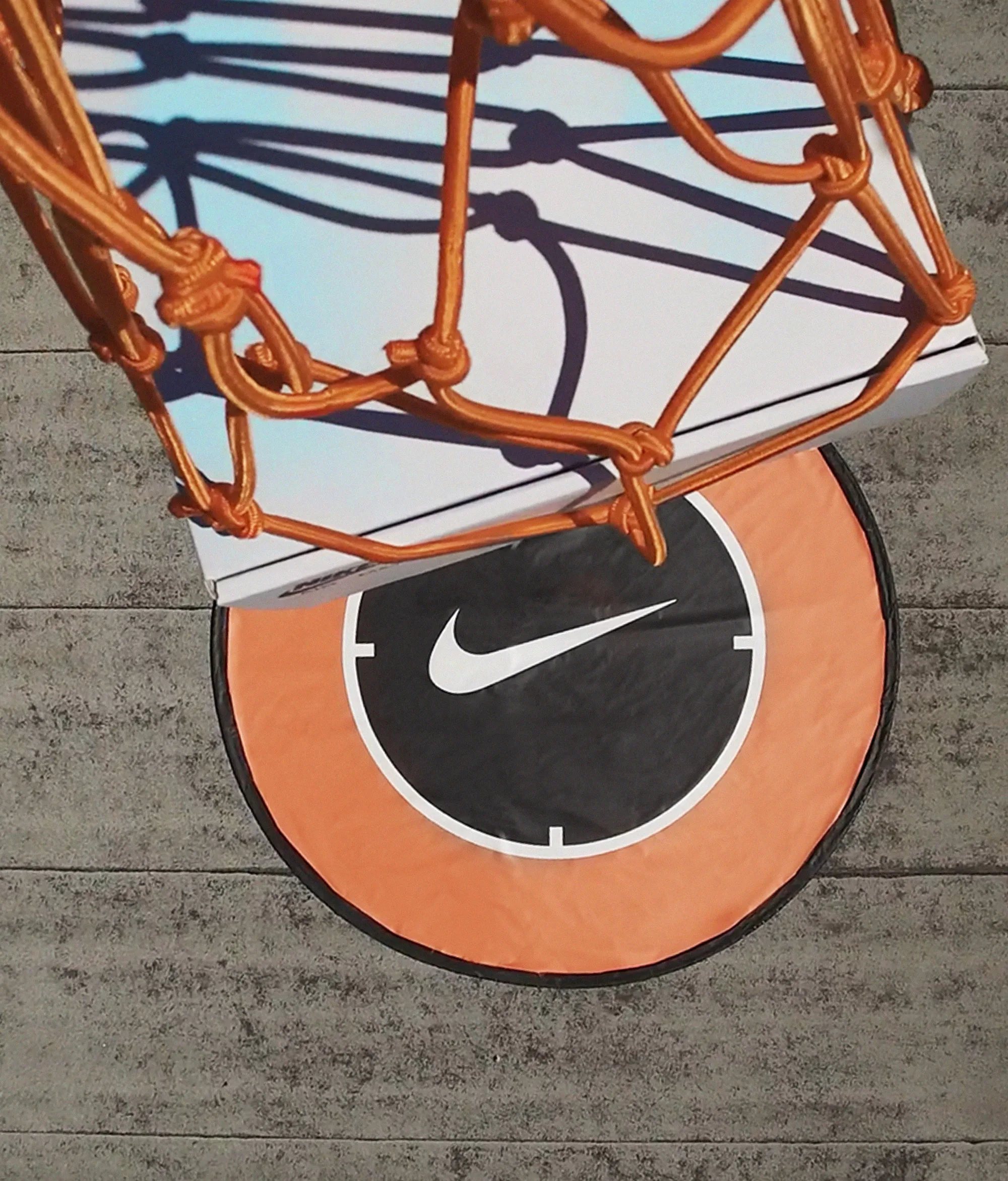 A close-up, top-down view of a basketball hoop with an orange net. A box is being dropped through the hoop, landing on a round orange and black mat with the white Nike logo. The surface beneath appears to be gray concrete.