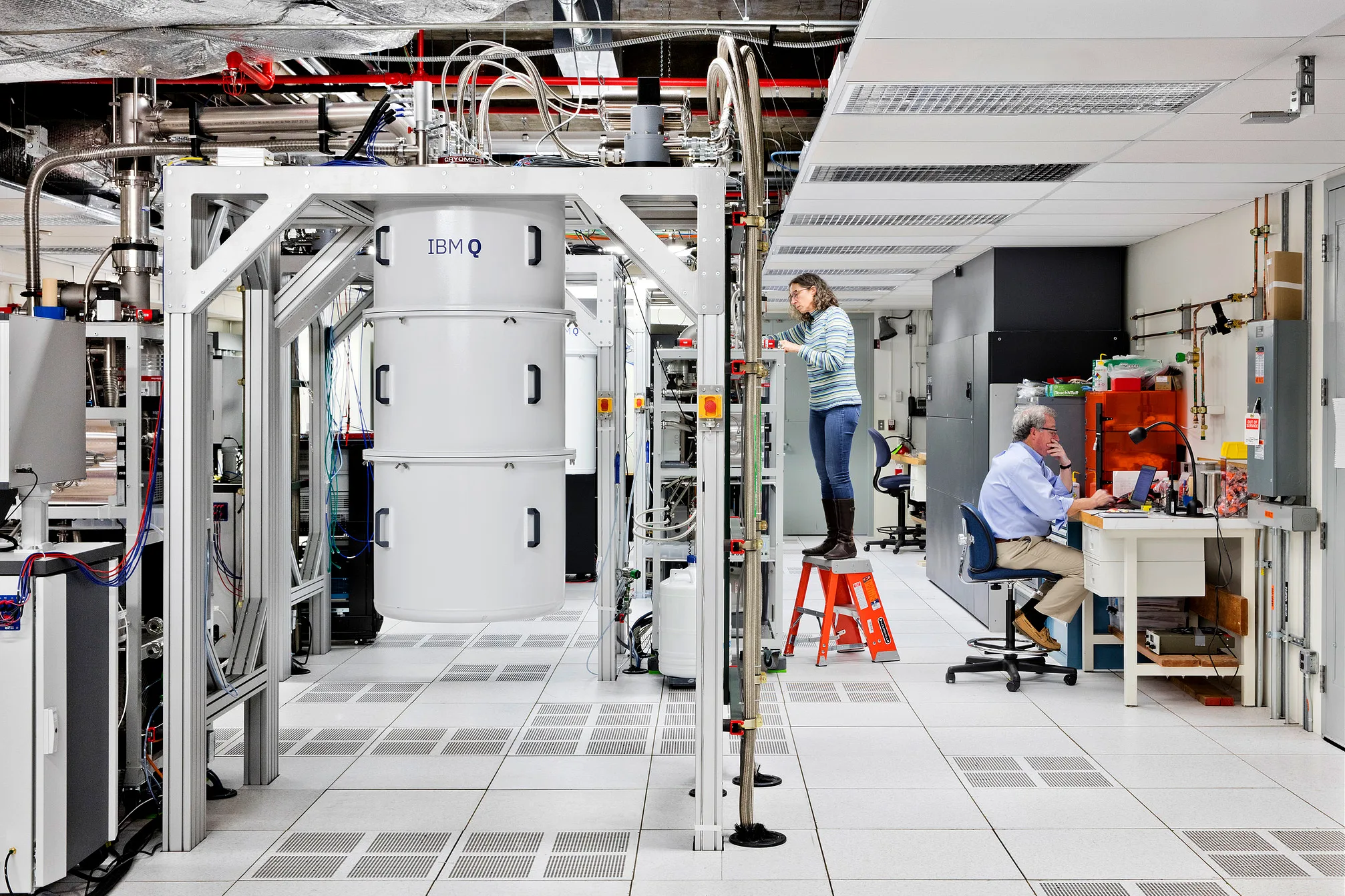 A modern laboratory with a large IBM Q quantum computer, a woman standing on a step stool working on equipment, and a man sitting at a desk using a computer. The environment is bright, clean, and technical.
