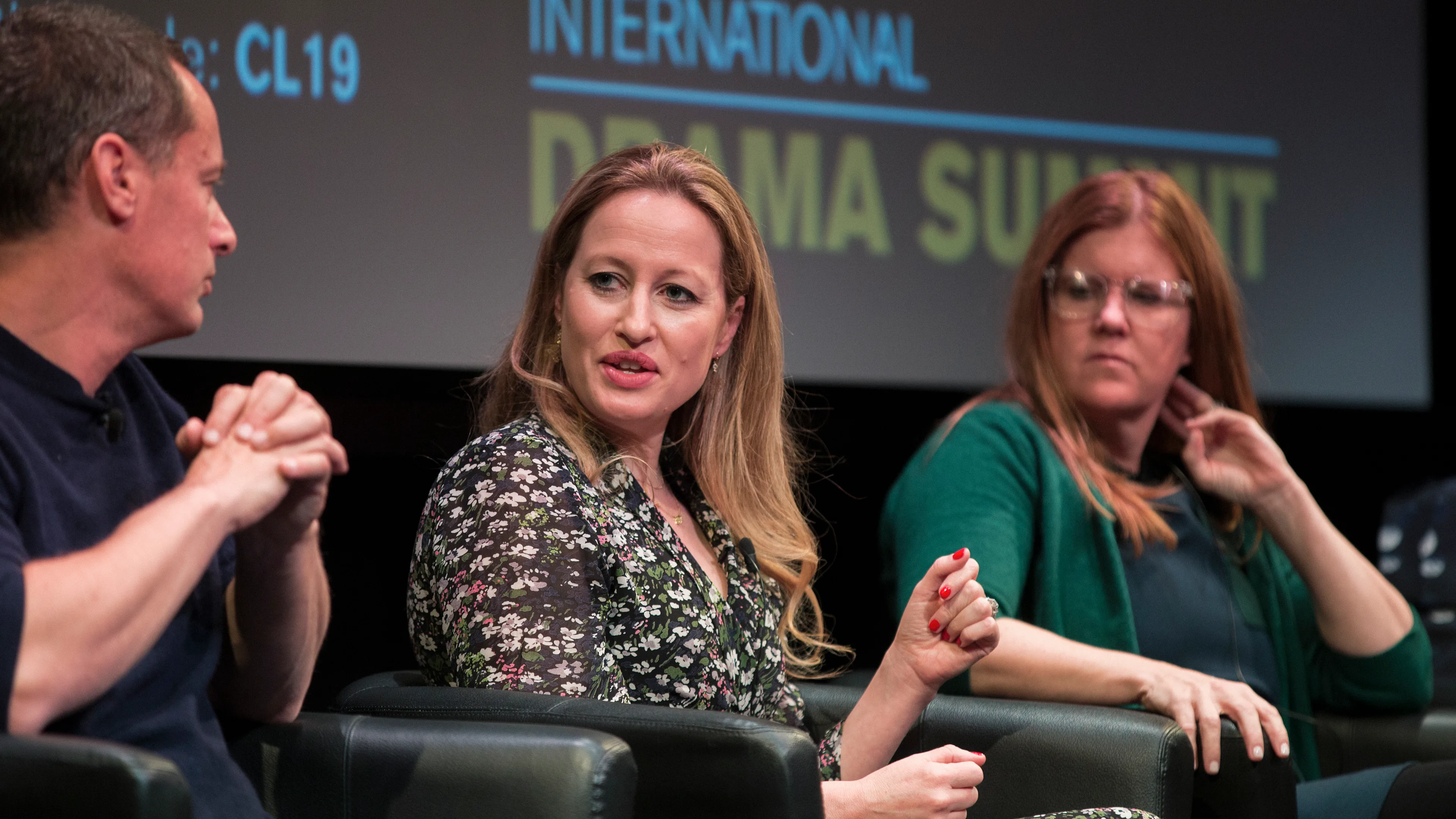 Three people sit on a panel in front of a screen that reads International Drama Summit. The woman in the center is speaking, while the other two listen attentively.
