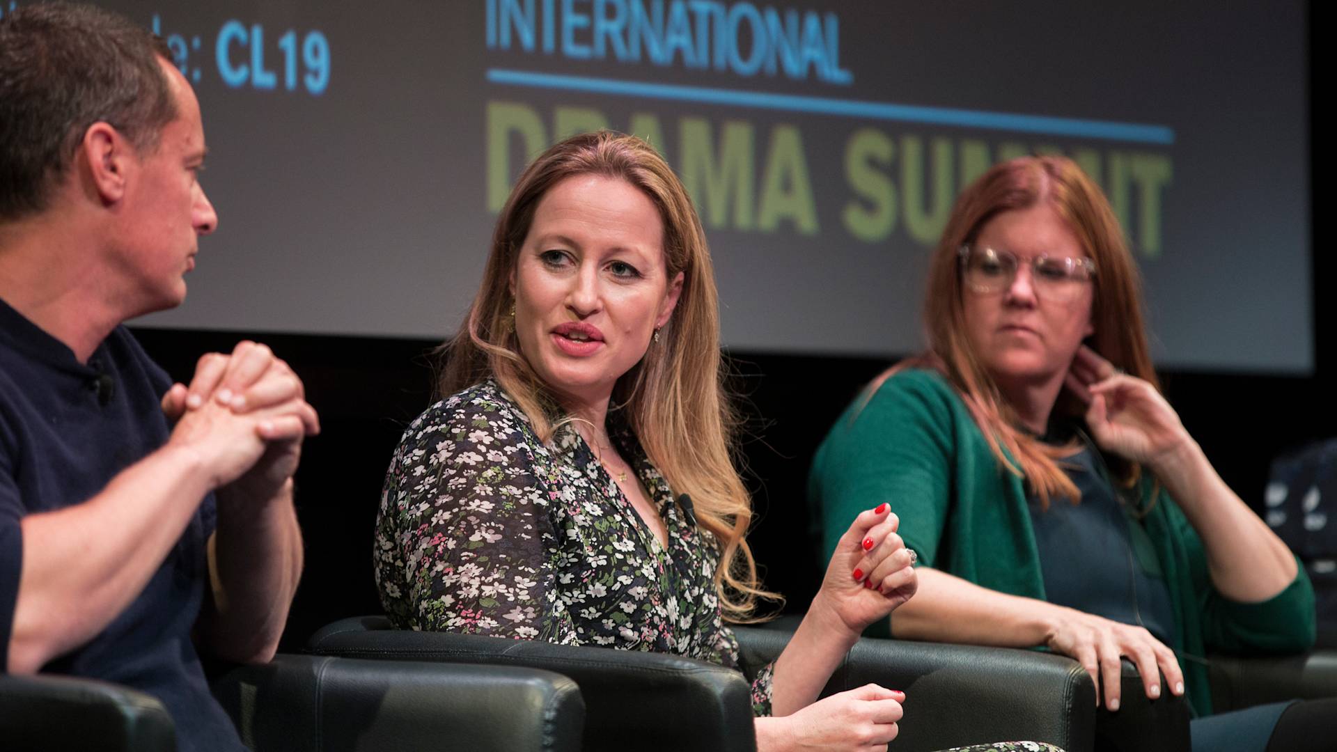 Three people sit on a panel in front of a screen that reads International Drama Summit. The woman in the center is speaking, while the other two listen attentively.