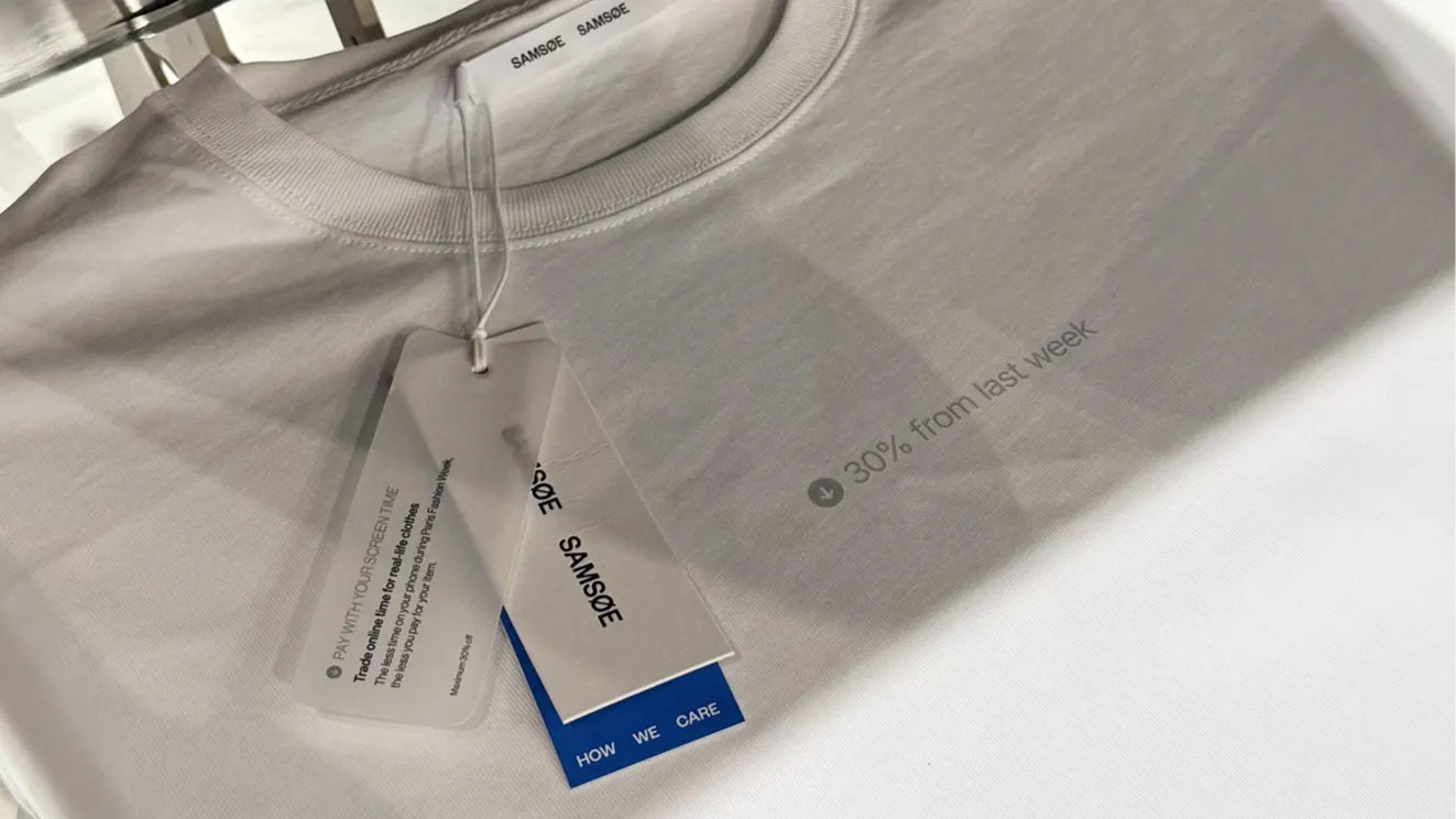 A white SAMSØE SAMSØE T-shirt with a subtle printed text 30% from last week and attached tags, one of which reads PAY WITH YOUR CONSCIENCE and another labeled HOW WE CARE.