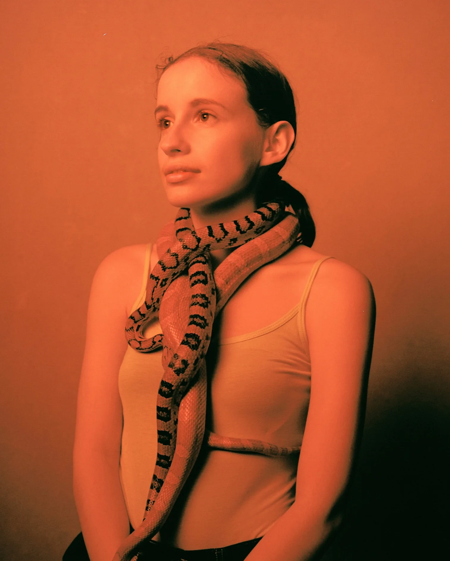 A woman with light skin and brown hair in a ponytail poses against a plain background, wearing a tank top. A large snake is draped around her neck and shoulders. The image has a reddish-orange tint.