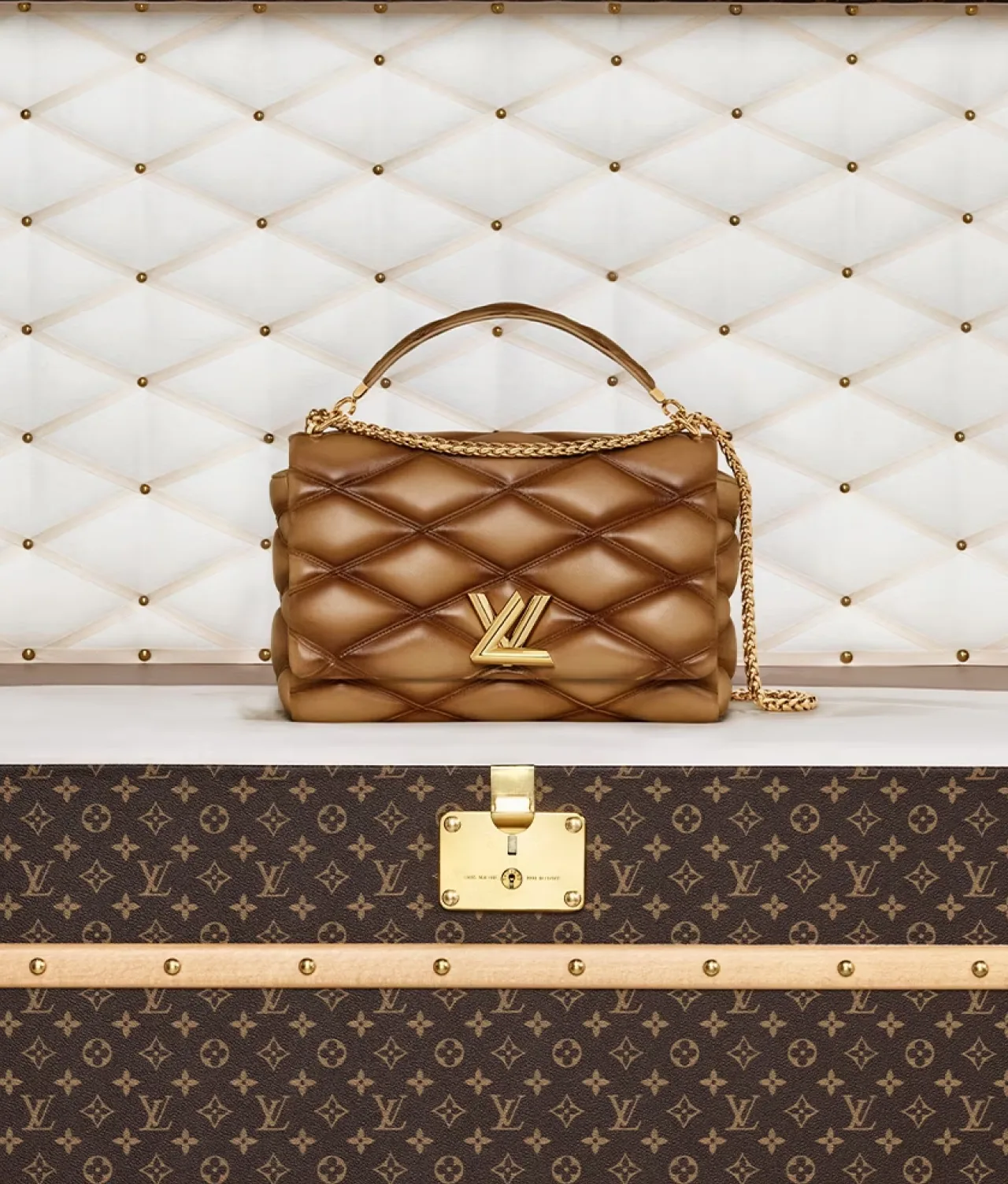 Brown quilted handbag with gold LV clasp. The bag is sitting on top of a large Louis Vuitton trunk with a white quilted background.