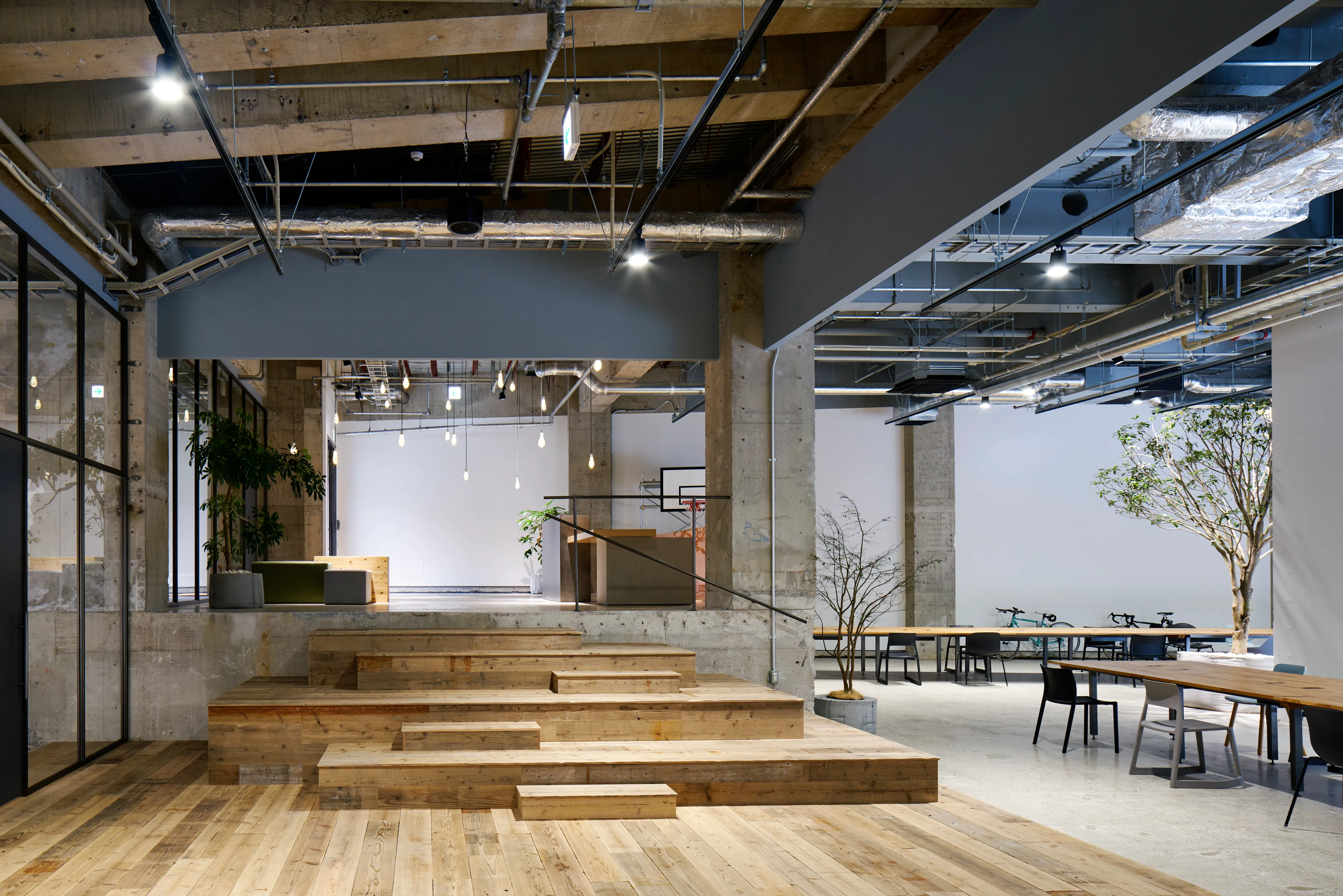 A modern, open-concept office space with exposed ceilings, wooden floors, tiered wooden seating, large tables, plants, and minimalist decor under bright overhead lighting.