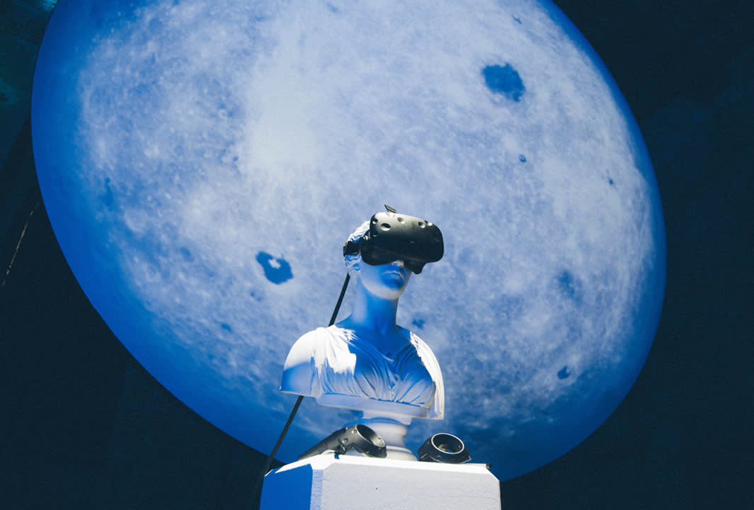 A white statue bust wearing a virtual reality headset is positioned in front of a large, illuminated image of the moon, creating a futuristic and surreal atmosphere.