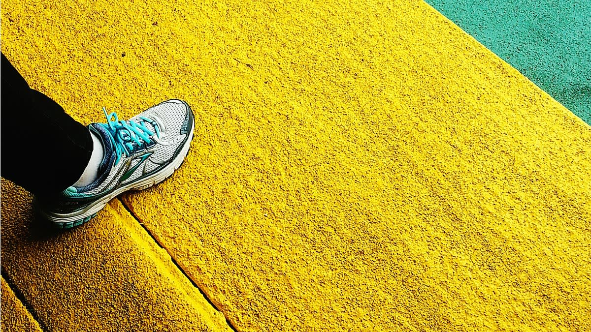 A person wearing a gray and blue sneaker steps onto a bright yellow textured surface, with green pavement visible in the background.