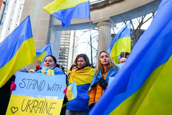 A group of people holding Ukrainian flags and a sign that reads Stand with Ukraine during a rally outdoors, with buildings and columns visible in the background.