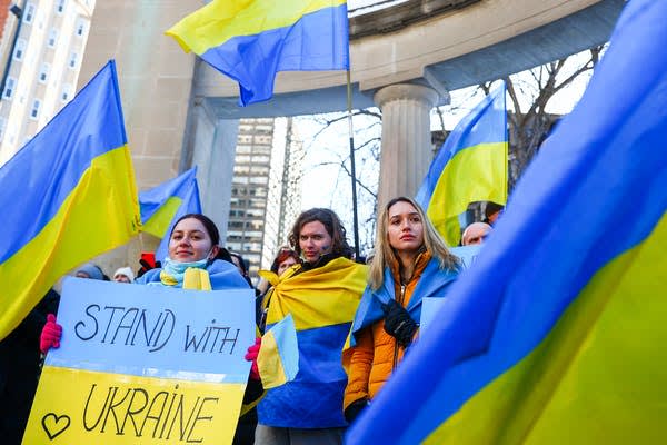 A group of people holding Ukrainian flags and a sign that reads Stand with Ukraine during a rally outdoors, with buildings and columns visible in the background.