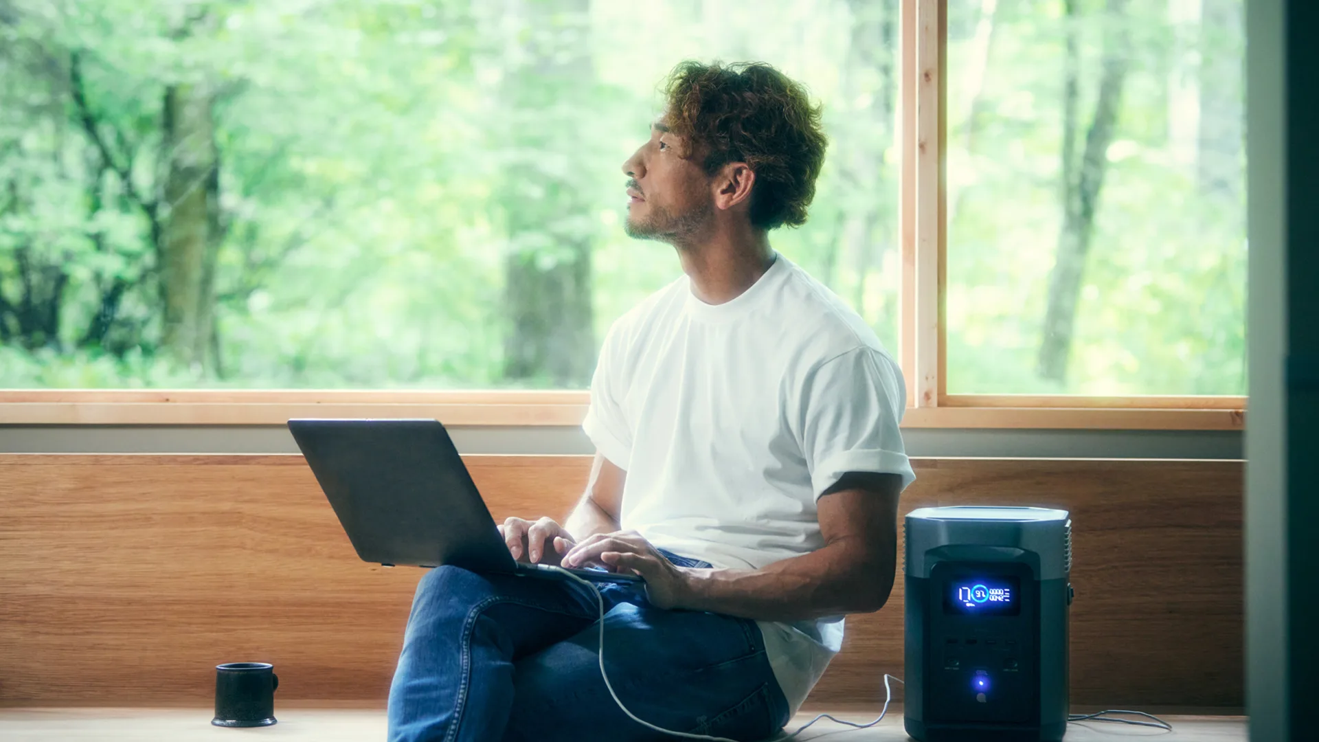 A man in a white t-shirt sits by a large window with a laptop on his lap, looking thoughtful. A portable power station connected to his laptop is on the bench beside him. Lush greenery is visible outside.