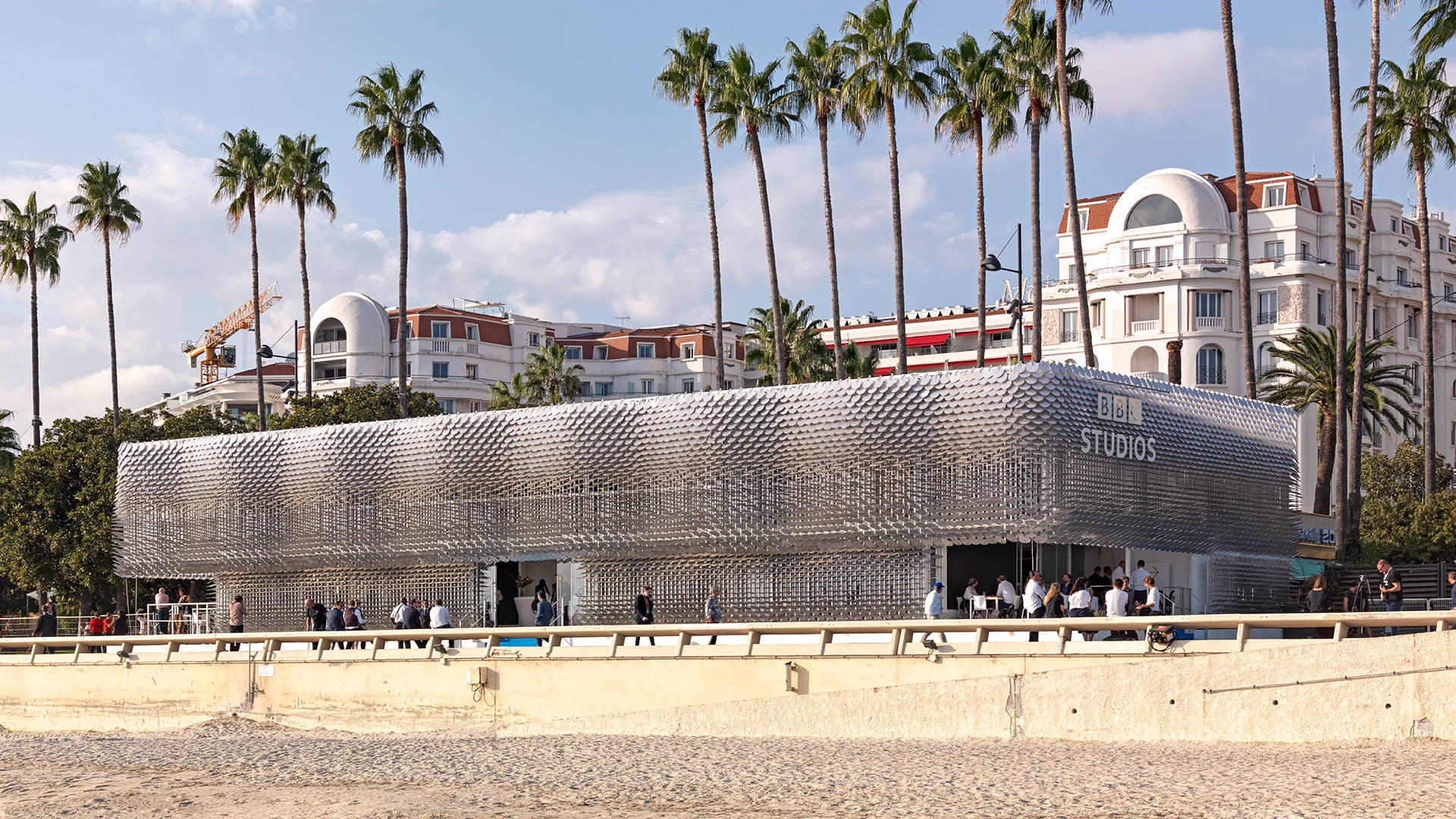 A modern, metallic building labeled “BNL Studios” sits on a beach promenade, surrounded by palm trees, with classic white buildings in the background and people walking nearby.