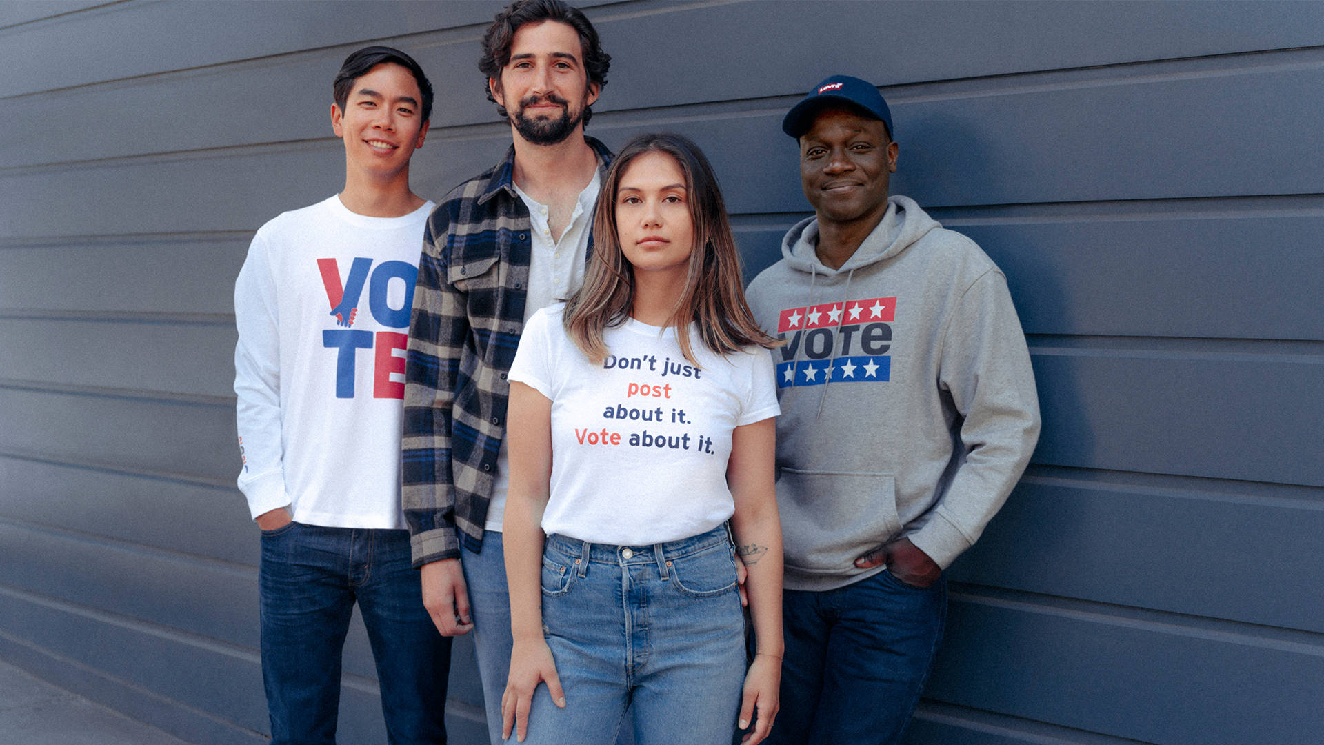 Four people stand in front of a gray wall, wearing casual clothes with slogans encouraging voting. Three wear shirts or hoodies that say Vote, and one wears a shirt that says, Dont just post about it. Vote about it.