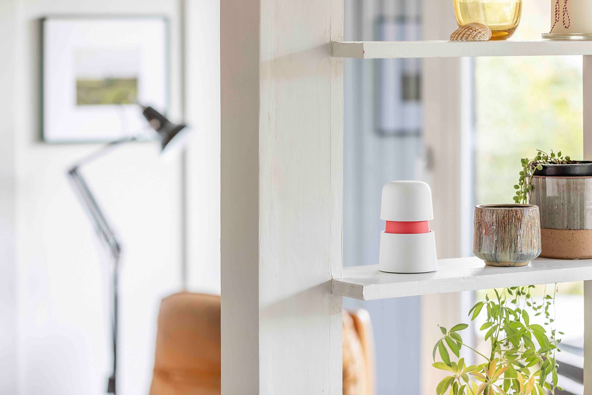 A white shelf with a small white device with a red band, a potted plant, and a mug. In the background, a blurred room with a lamp, framed art, and a chair is visible.