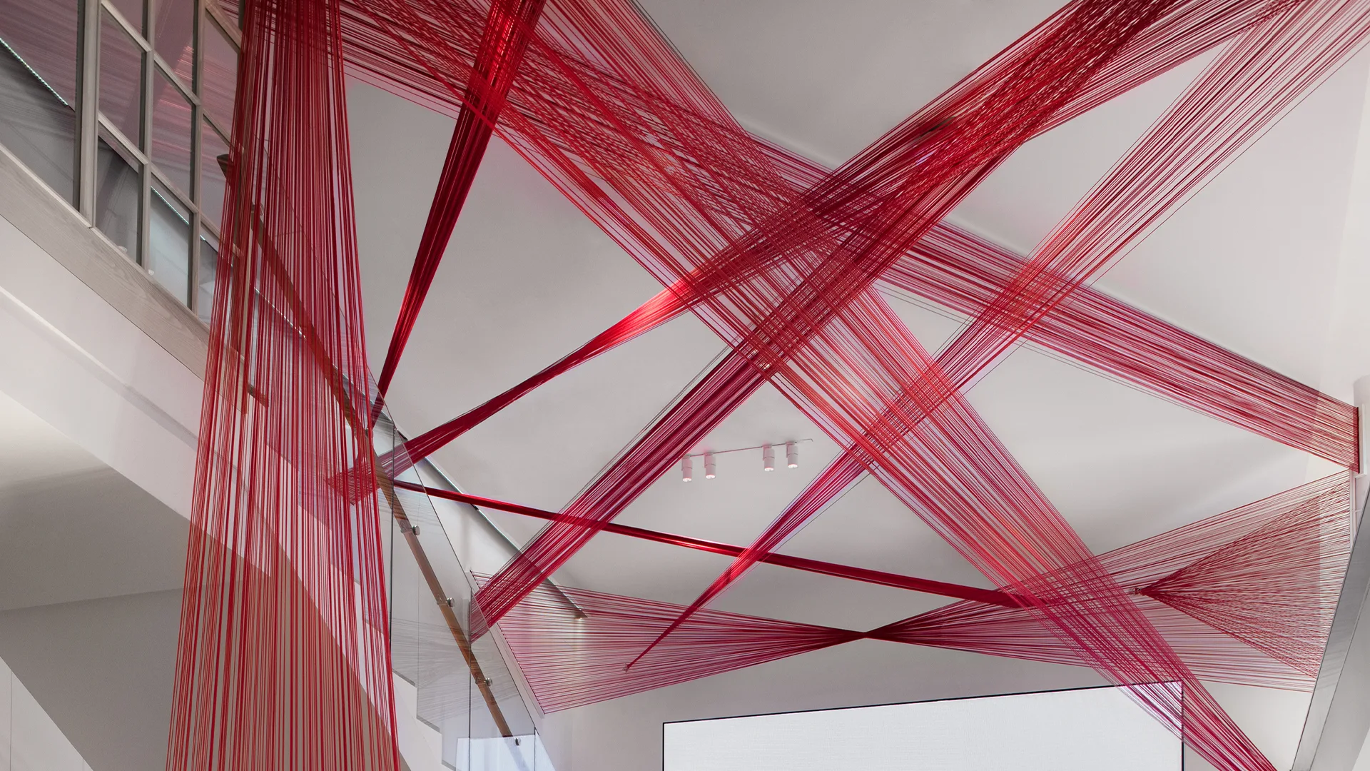 Red threads are stretched in intricate, overlapping patterns across a white stairwell, creating a geometric web-like art installation suspended between the railings and ceiling.
