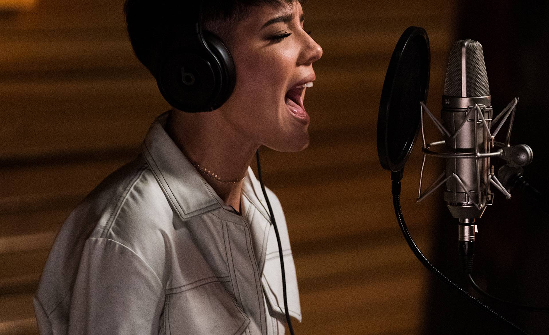 A person wearing headphones and a light-colored shirt passionately sings into a professional microphone in a recording studio.
