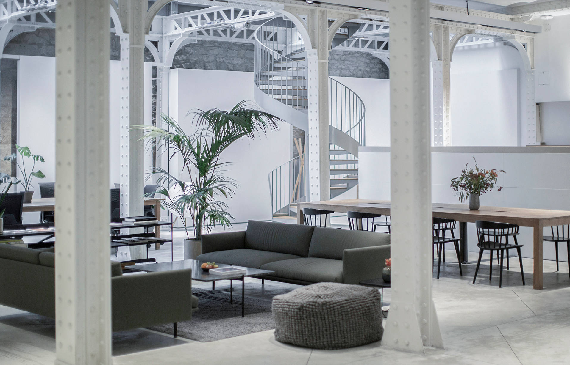 Modern open-plan office featuring metal beams, green sofas, a large wooden table with chairs, indoor plants, and a spiral staircase in the background. The space is bright with industrial and minimalist decor.