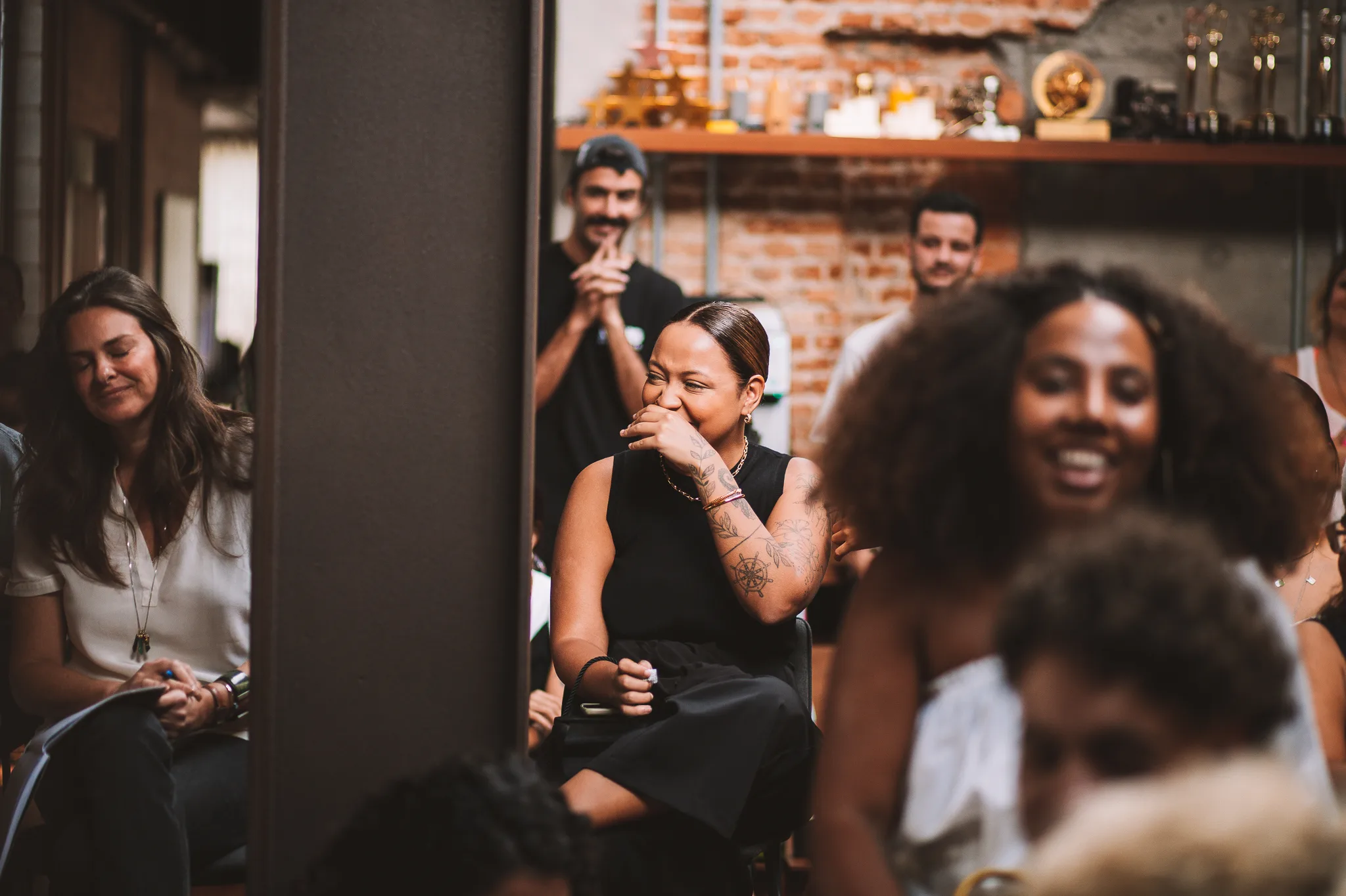 A group of people sitting indoors, smiling and laughing. The focus is on a woman in black with tattoos, covering her mouth while laughing. The background shows shelves with trophies and exposed brick walls.