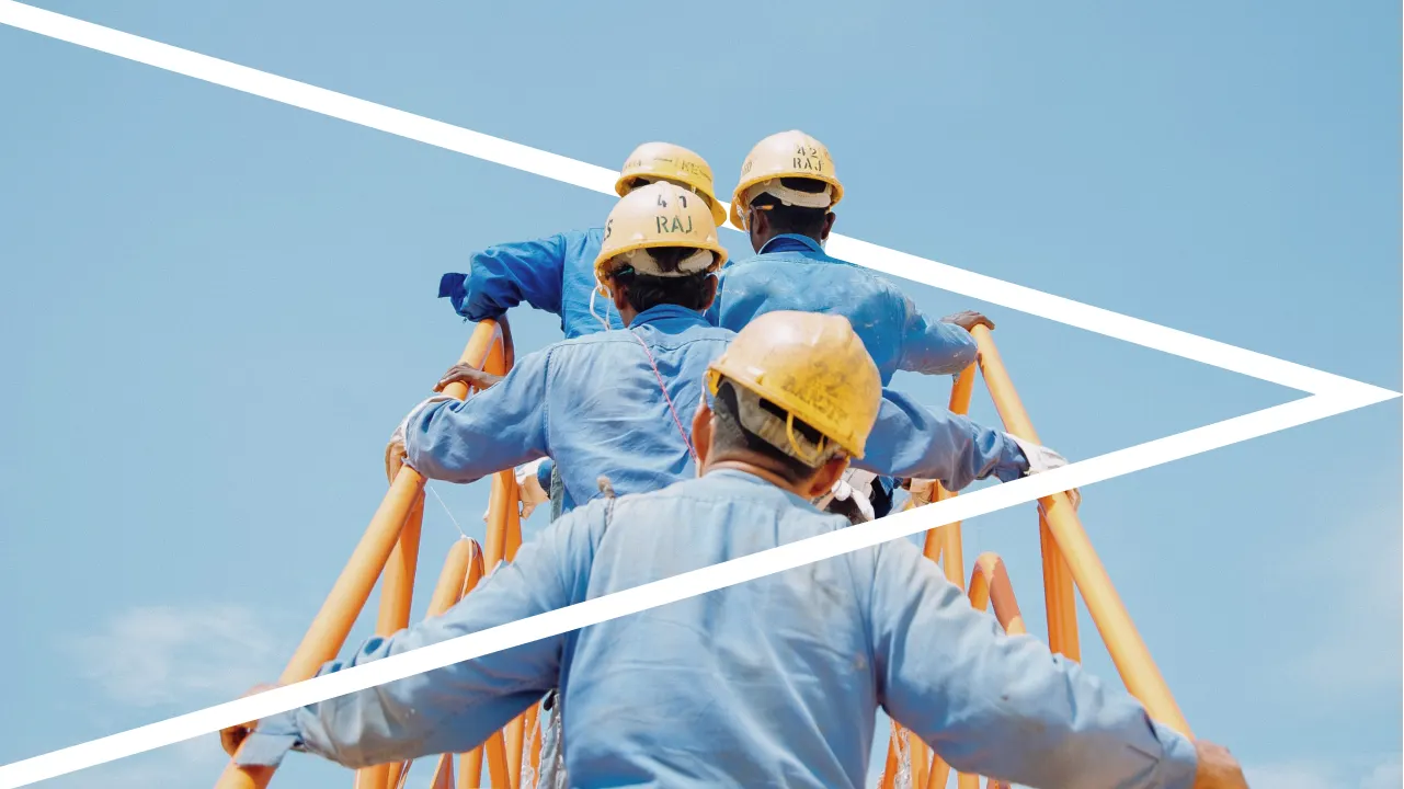 Several construction workers in blue uniforms and yellow helmets climb a yellow metal structure under a clear blue sky, with two diagonal white lines crossing the image.