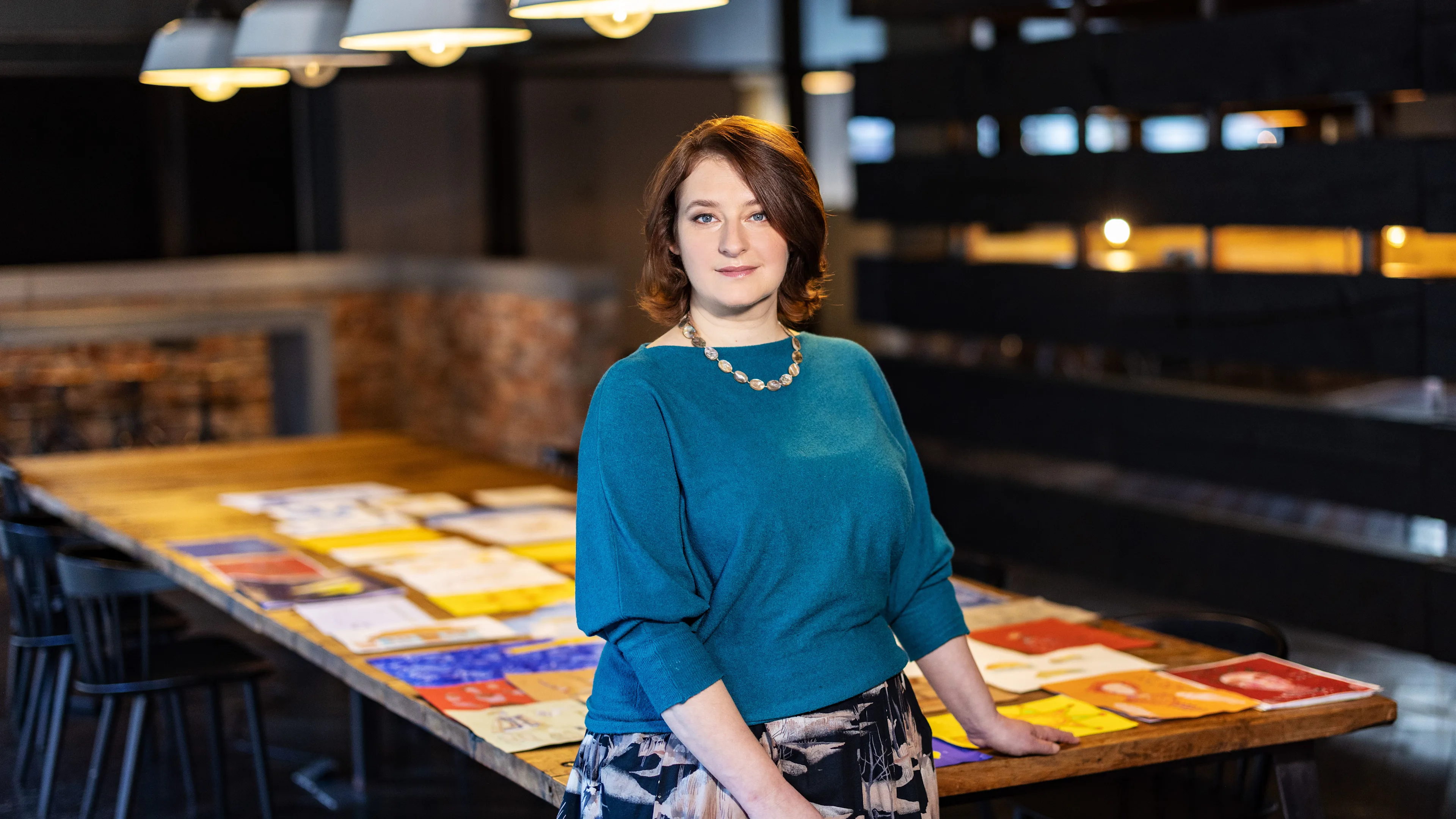 A woman with shoulder-length brown hair, wearing a blue top and patterned skirt, stands beside a long wooden table covered with colorful papers and artworks in a modern, dimly lit room.