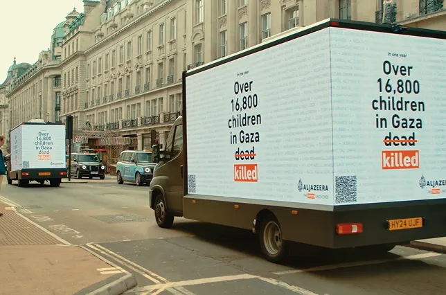 Two vans drive along a city street, each displaying a billboard reading, In one year: Over 16,800 children in Gaza killed, with the words dead crossed out and replaced with killed.
